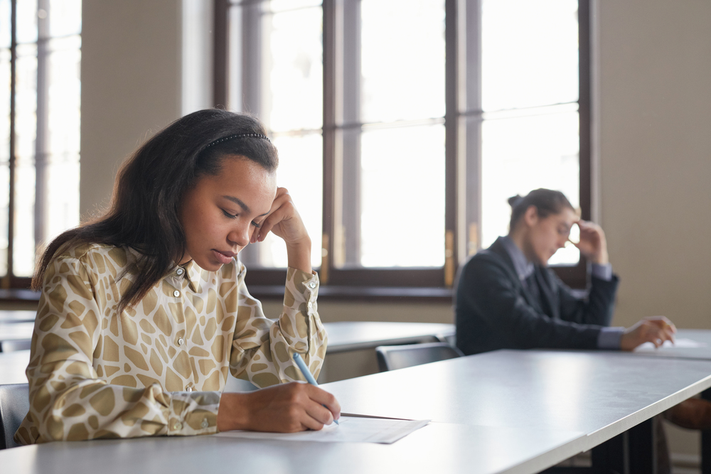 Side view portrait of two students taking exam in row while sitting at desk with social distancing, focus on young African-American woman in foreground, copy space