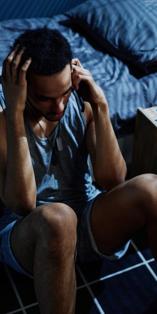 Guy with post traumatic syndrome calling 911 to get psychological help or consultation of specialist while sitting by bed on the floor